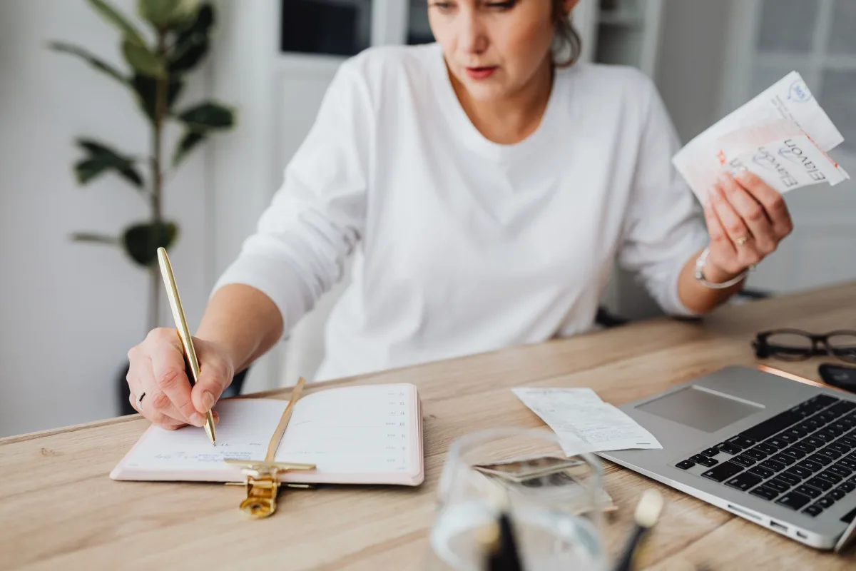 A woman writing in a notebook at a desk with a laptop, receipts and a calculator