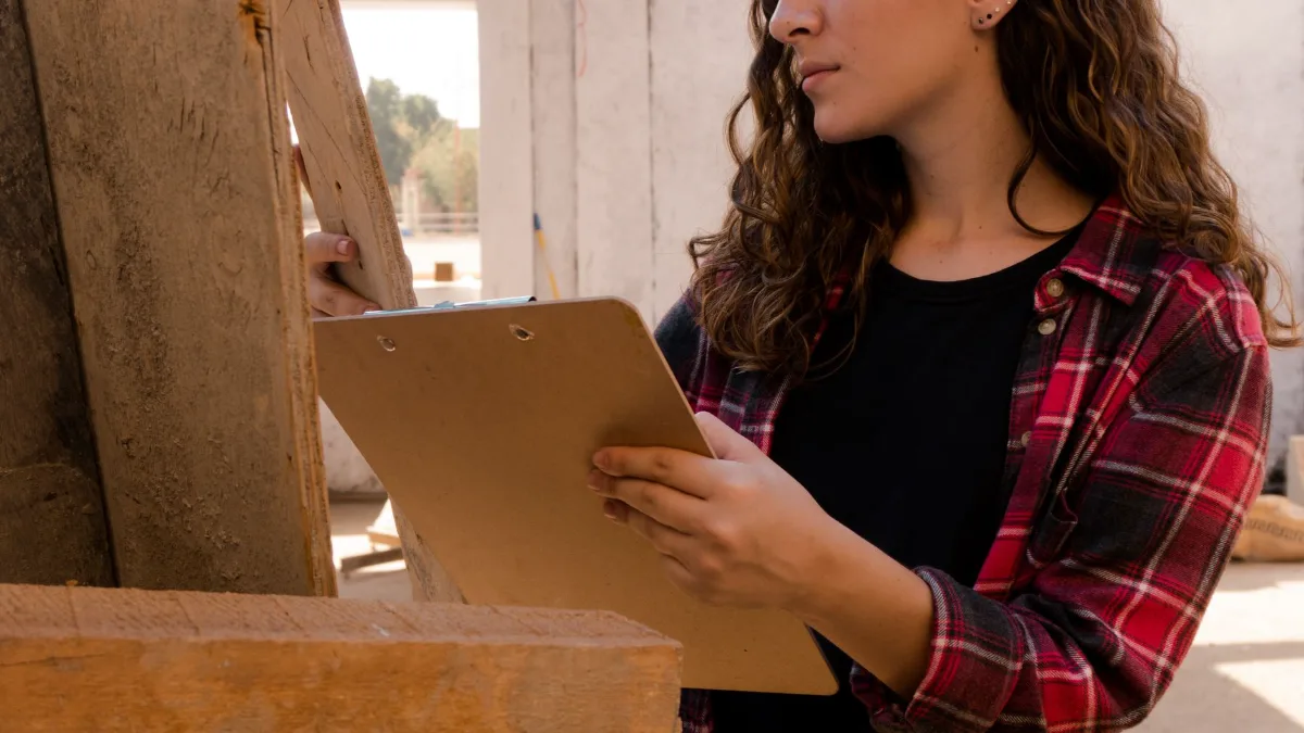 A contractor in a plaid shirt holding a clipboard and inspecting wooden planks